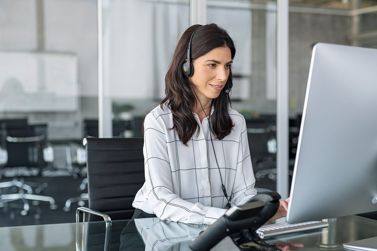 Mature secretary working in call center with headset. Confident telephone operator in modern office working on computer while taking calls. Smiling customer support operator at work with copy space.