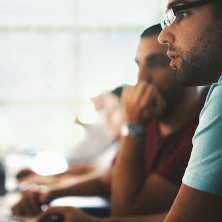 Side view of two partially unrecognizable men sitting in front of a computer.They are looking at computer codes on the screen and thinking of how to do their task. Blurry unrecognizable people in background.
